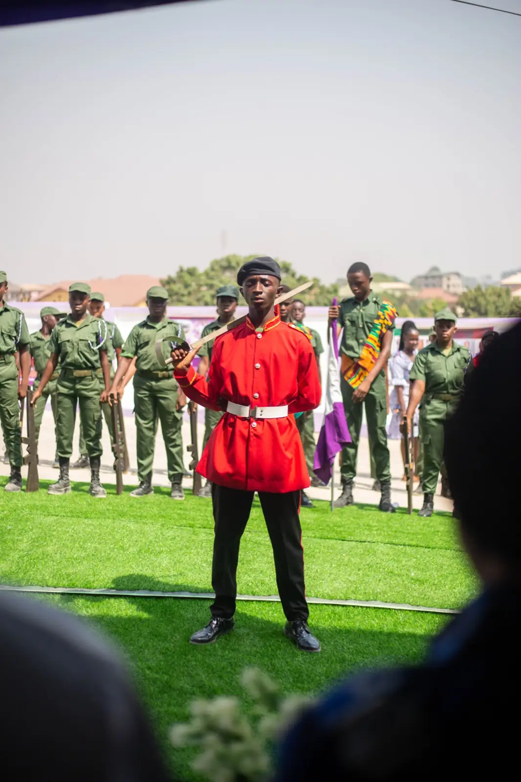 A pupil in a red ceremonial tunic leading the school parade on the field